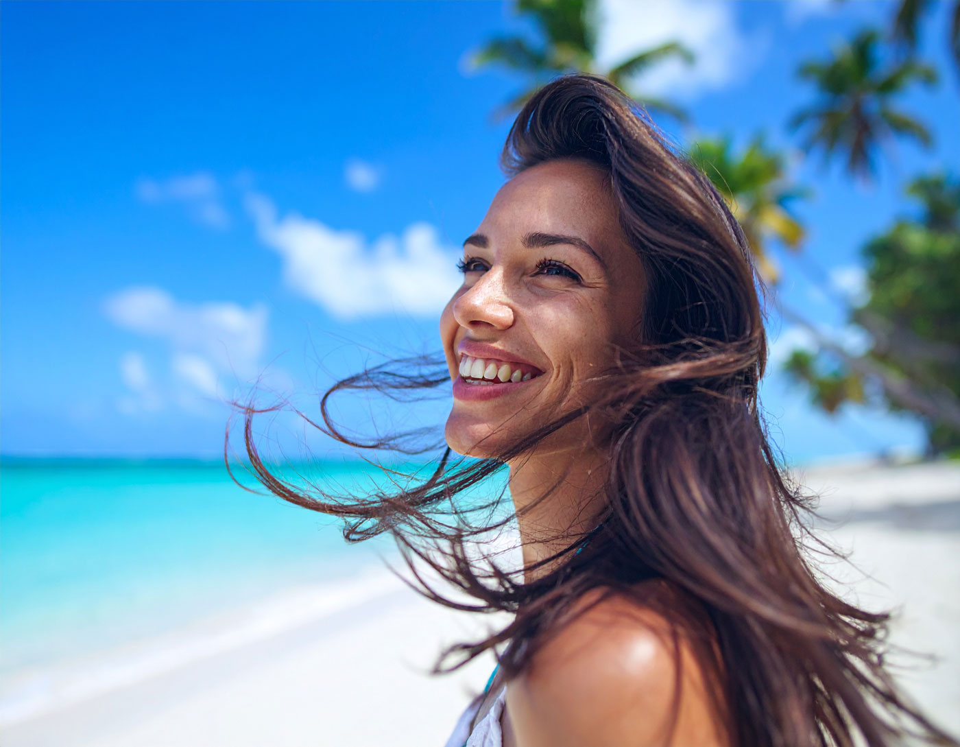 Nahaufnahme einer lachenden Frau mit langen, glänzenden Echthaar-Extensions am weißen Sandstrand, im Hintergrund türkisblaues Meer und Palmen unter blauem Himmel.